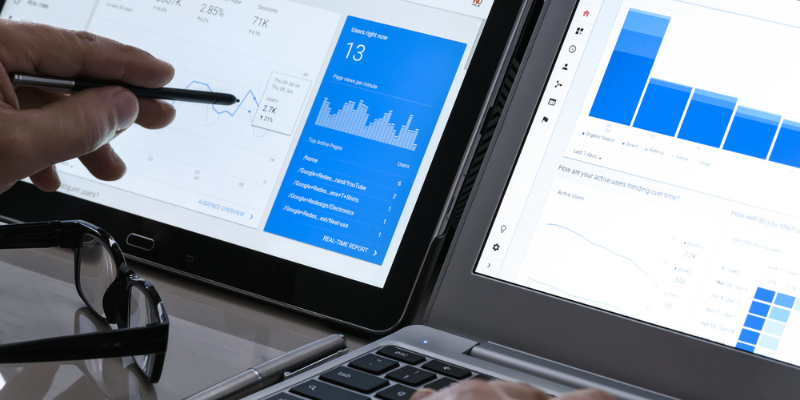 Someone reviewing data on two screens with glasses sitting down on the table in front of the screen.